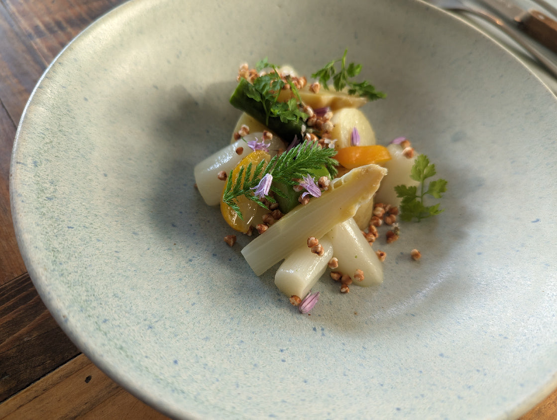 Assiette gastronomique composée d'asperges blanches et vertes, garnie de fines herbes, de fleurs comestibles et de graines de sarrasin grillées.