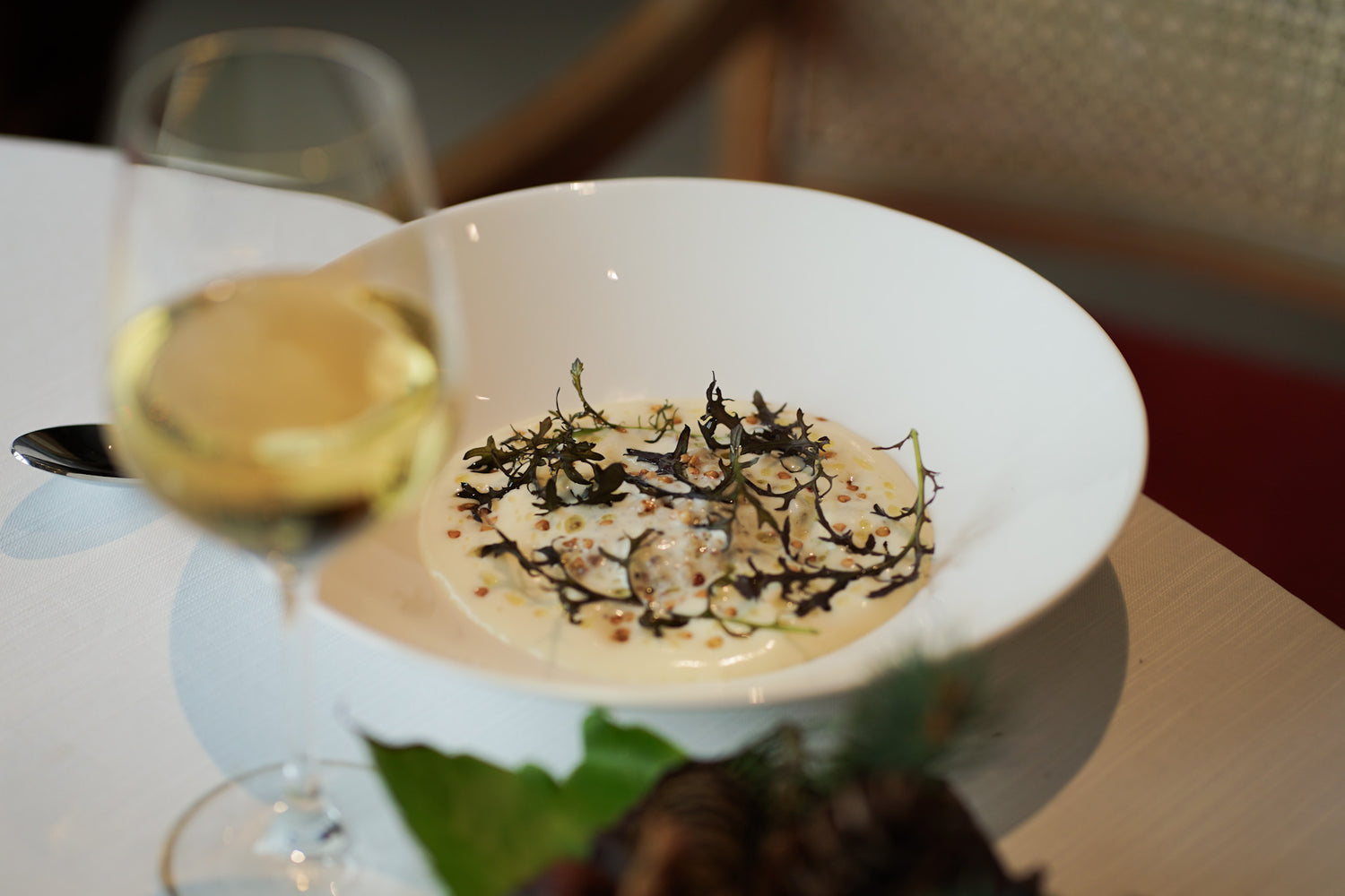 Plat gastronomique raffiné servi dans une assiette blanche profonde, composé d'une sauce crémeuse, de garnitures croustillantes et d'herbes aromatiques, accompagné d'un verre de vin blanc en arrière-plan sur une table élégamment dressée.