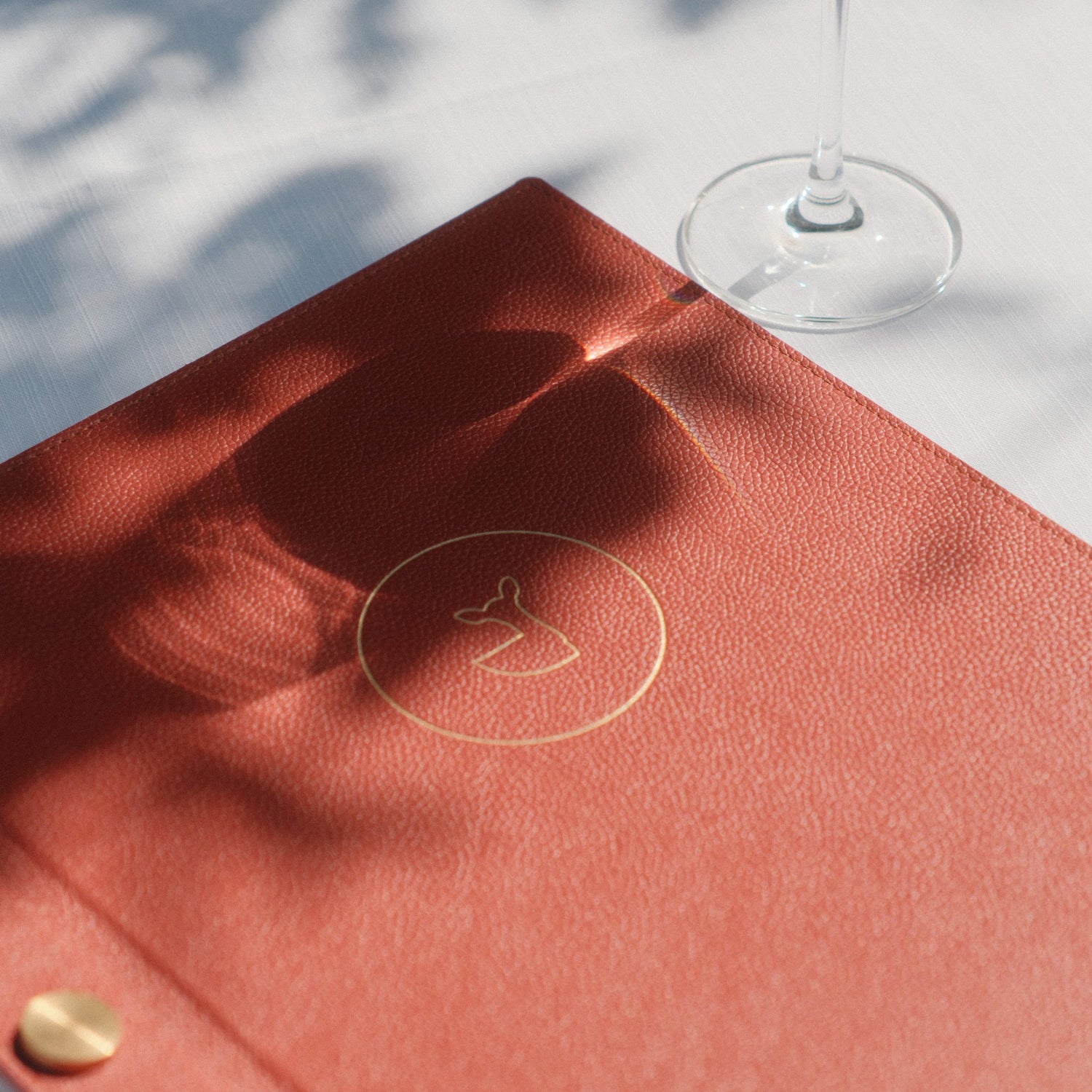 Menu de restaurant en cuir rouge avec un logo doré en relief, posé sur une table élégante avec un verre de vin blanc, sous un jeu d'ombres naturelles projetées par la lumière.