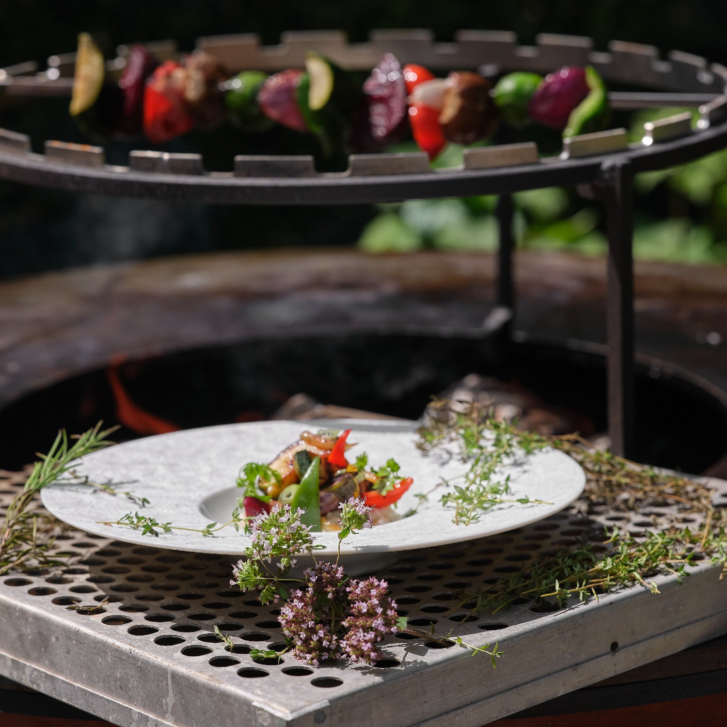 Assiette de légumes grillés présentée devant un barbecue avec des brochettes de légumes en train de cuire, décorée de plantes aromatiques.