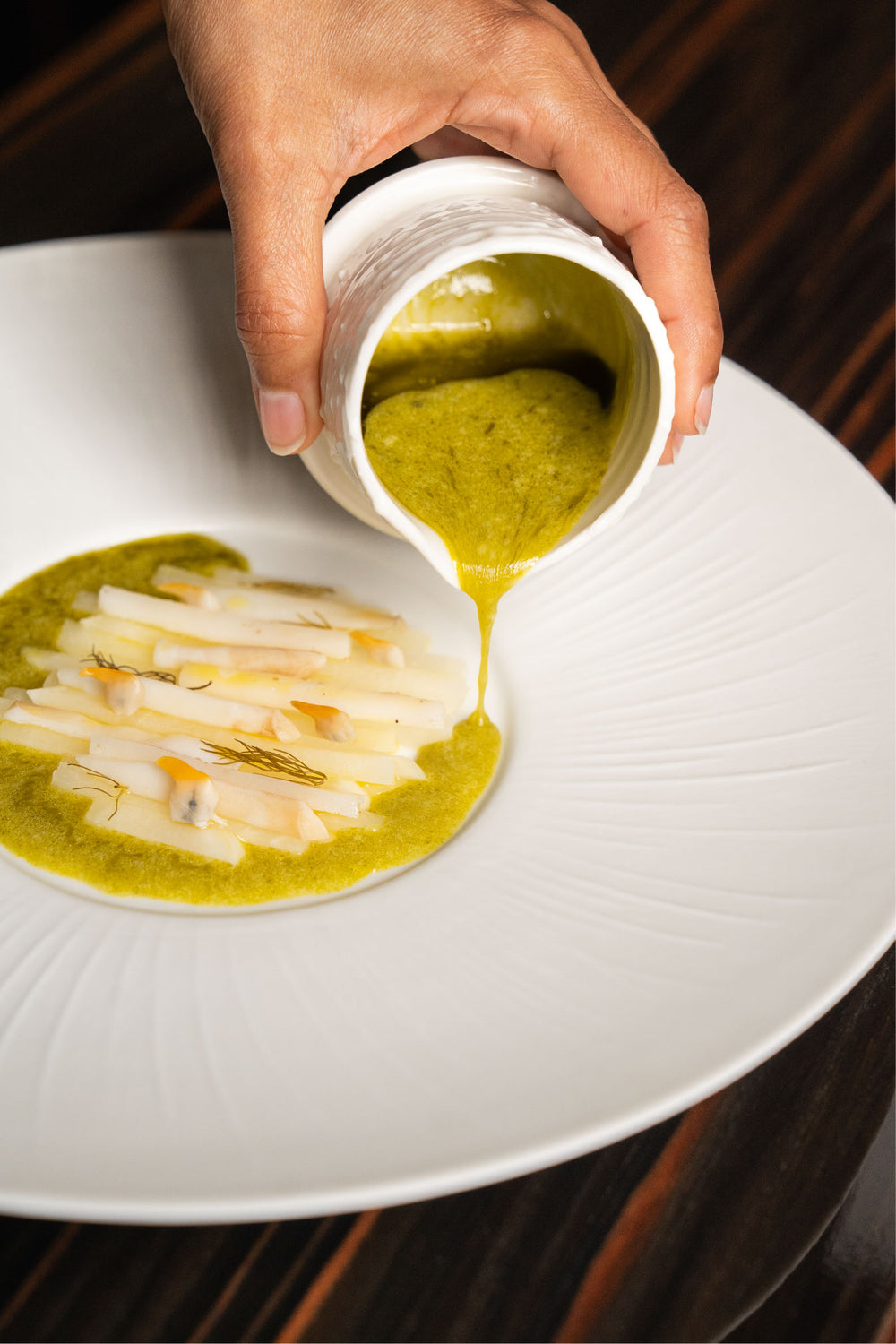 Plat gastronomique d’asperges et de sauce verte versée à la main, dressé avec précision dans une assiette blanche.