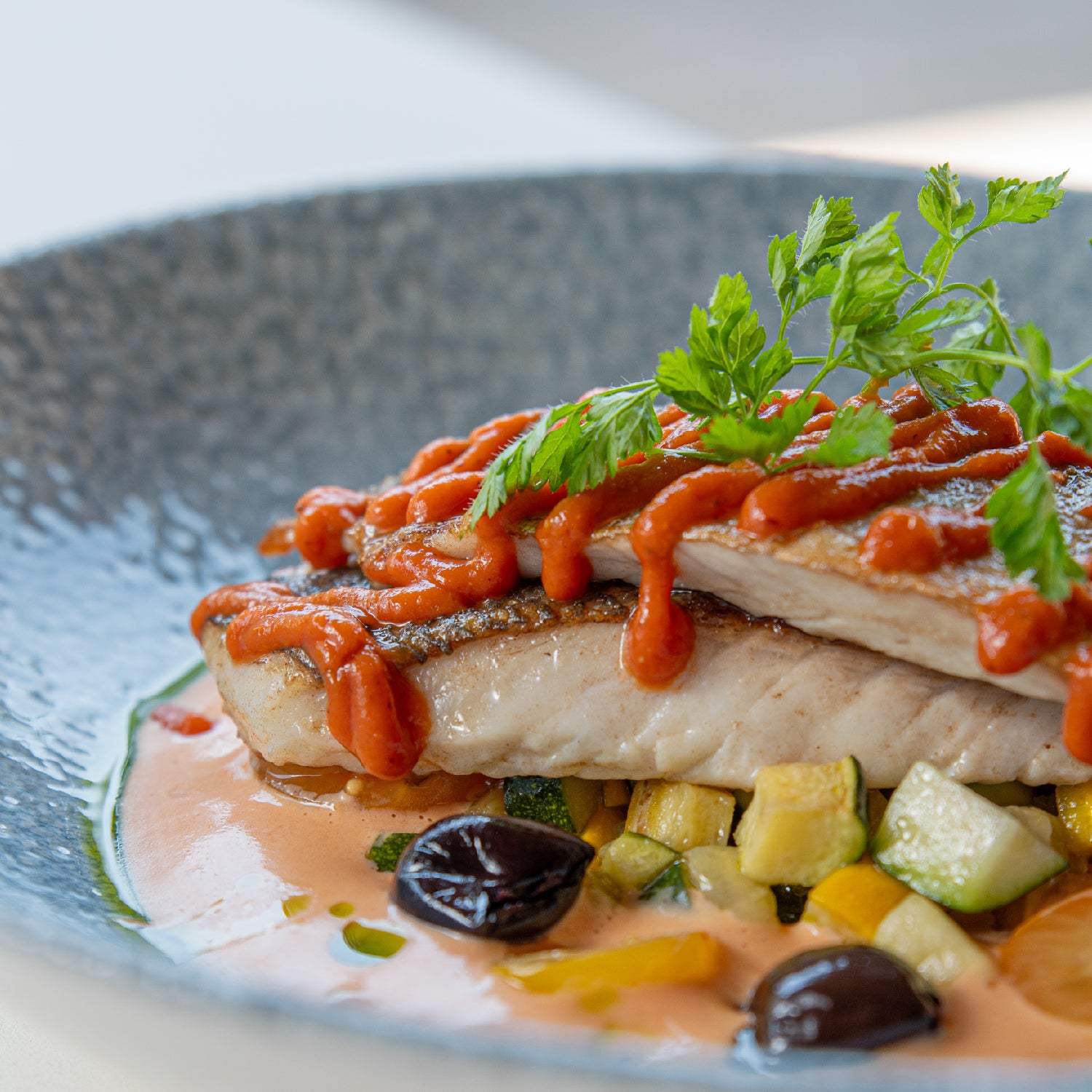 Poisson grillé nappé de sauce rouge, accompagné de légumes d’été et olives noires, présenté dans une assiette bleu texturée avec une touche d’herbes fraîches.