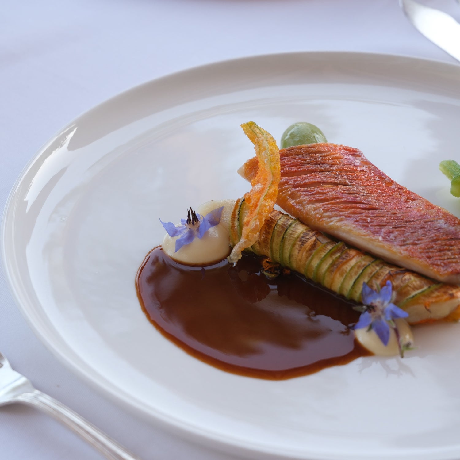 Filet de rouget accompagné de légumes finement taillés, fleurs comestibles et sauce brune dressée avec précision sur une assiette blanche.