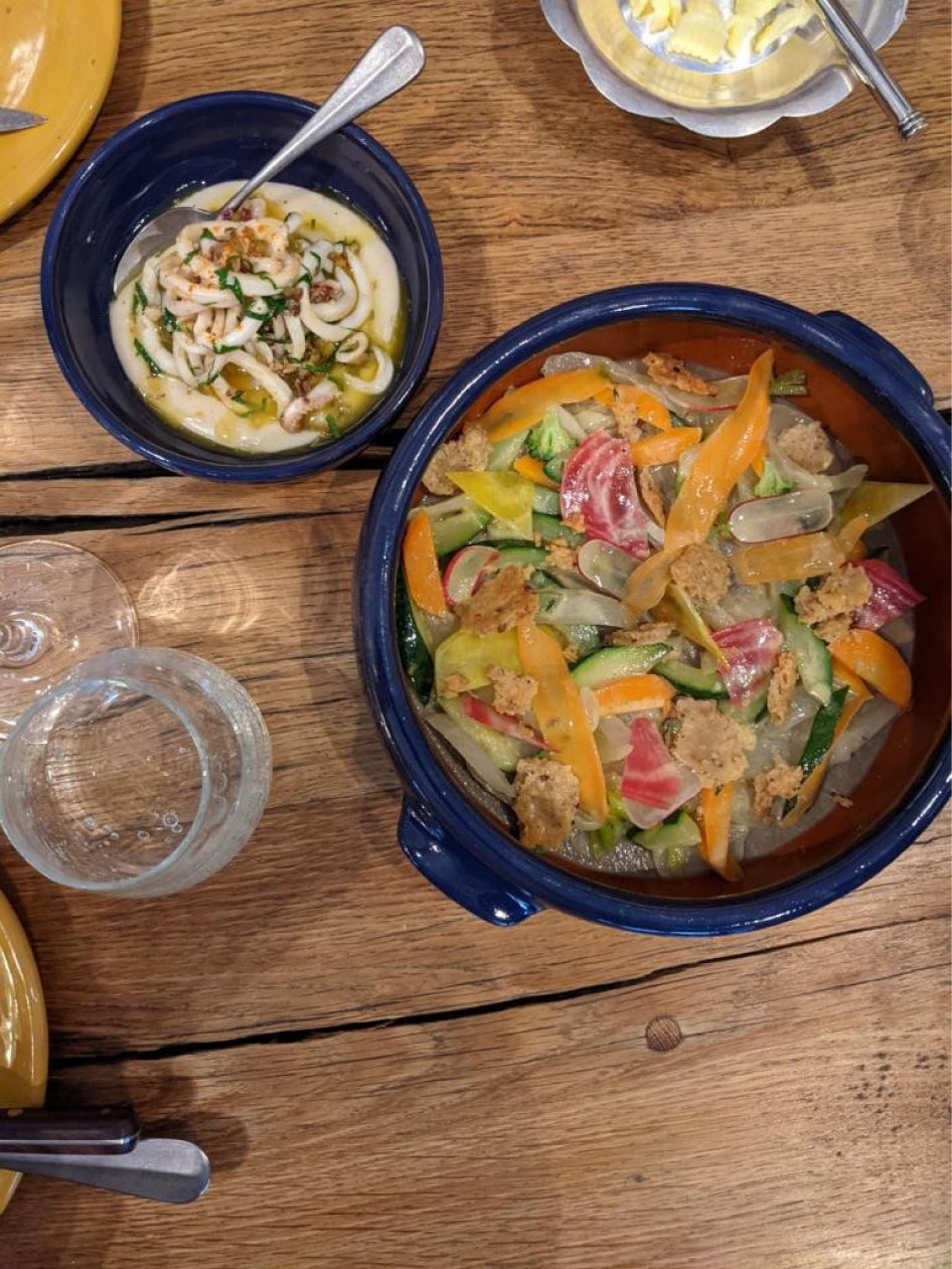 Repas coloré avec des légumes frais dans un bol bleu, accompagné d'un plat de calamars dans une sauce onctueuse et du beurre servi dans un bol en métal, le tout sur une table en bois rustique.