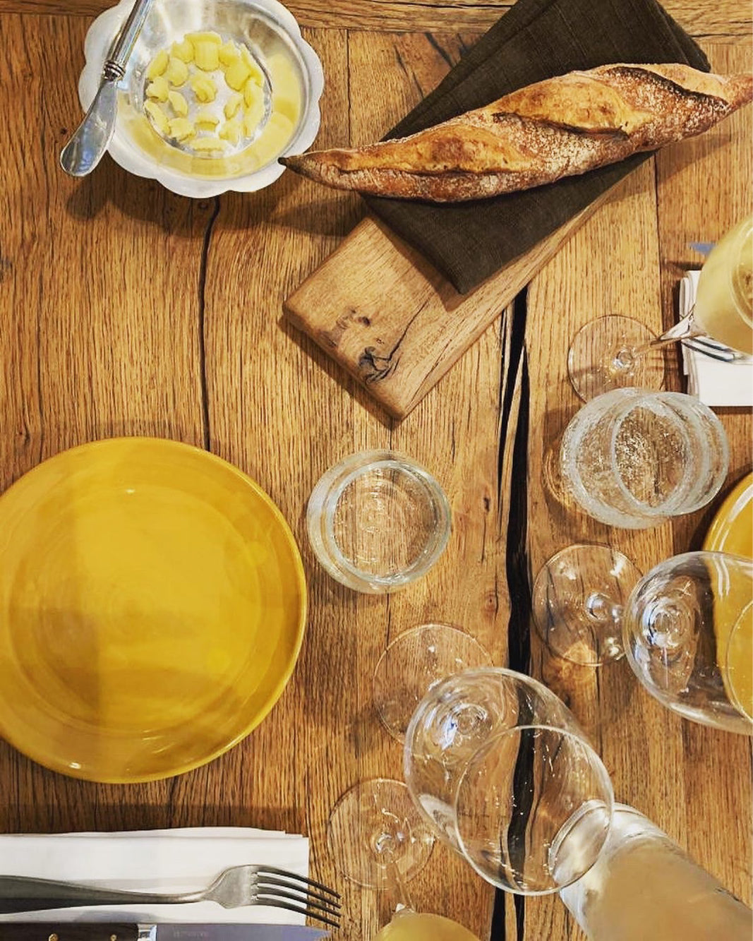 Table rustique en bois avec une baguette croustillante, une assiette jaune, du beurre présenté dans un bol en métal et des verres élégants, prête pour un repas convivial.