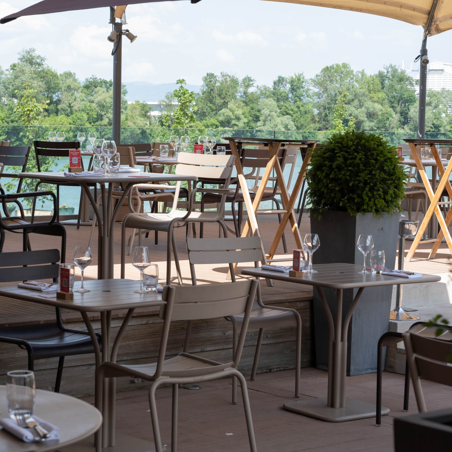 Terrasse de restaurant élégante avec vue sur une rivière bordée de verdure, tables dressées avec verrerie et ardoise de menu à l'entrée.