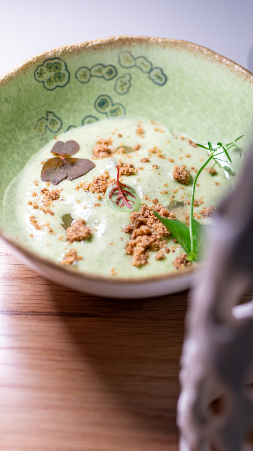 Un plat raffiné d'escargots, présenté dans une assiette en céramique verte. La préparation est agrémentée d'une mousse verte crémeuse, de miettes croquantes et de feuilles de décoration.