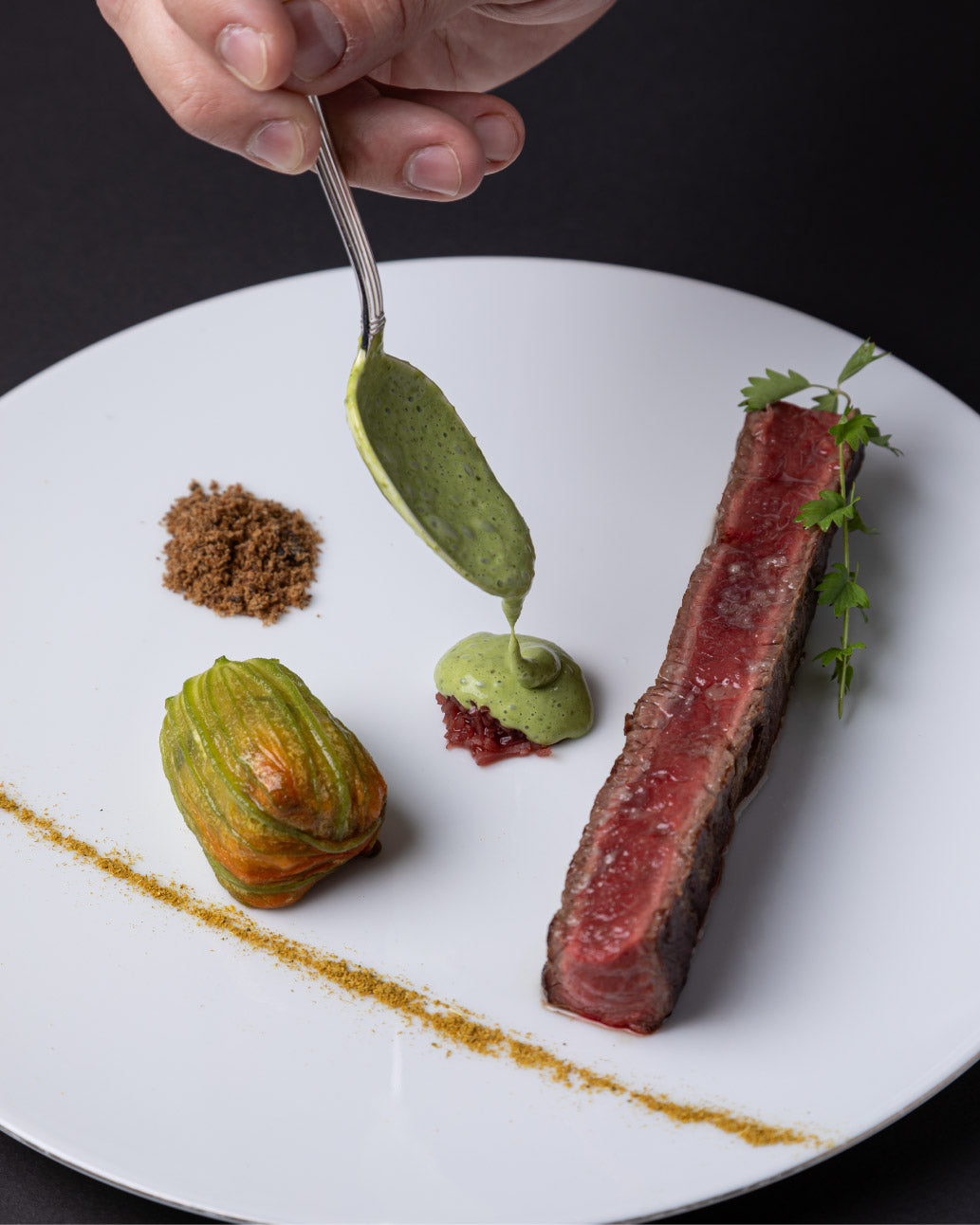 Plat gastronomique avec un morceau de viande saignante, une fleur de courgette farcie et une sauce verte délicatement placée sur l'assiette, le tout joliment présenté.