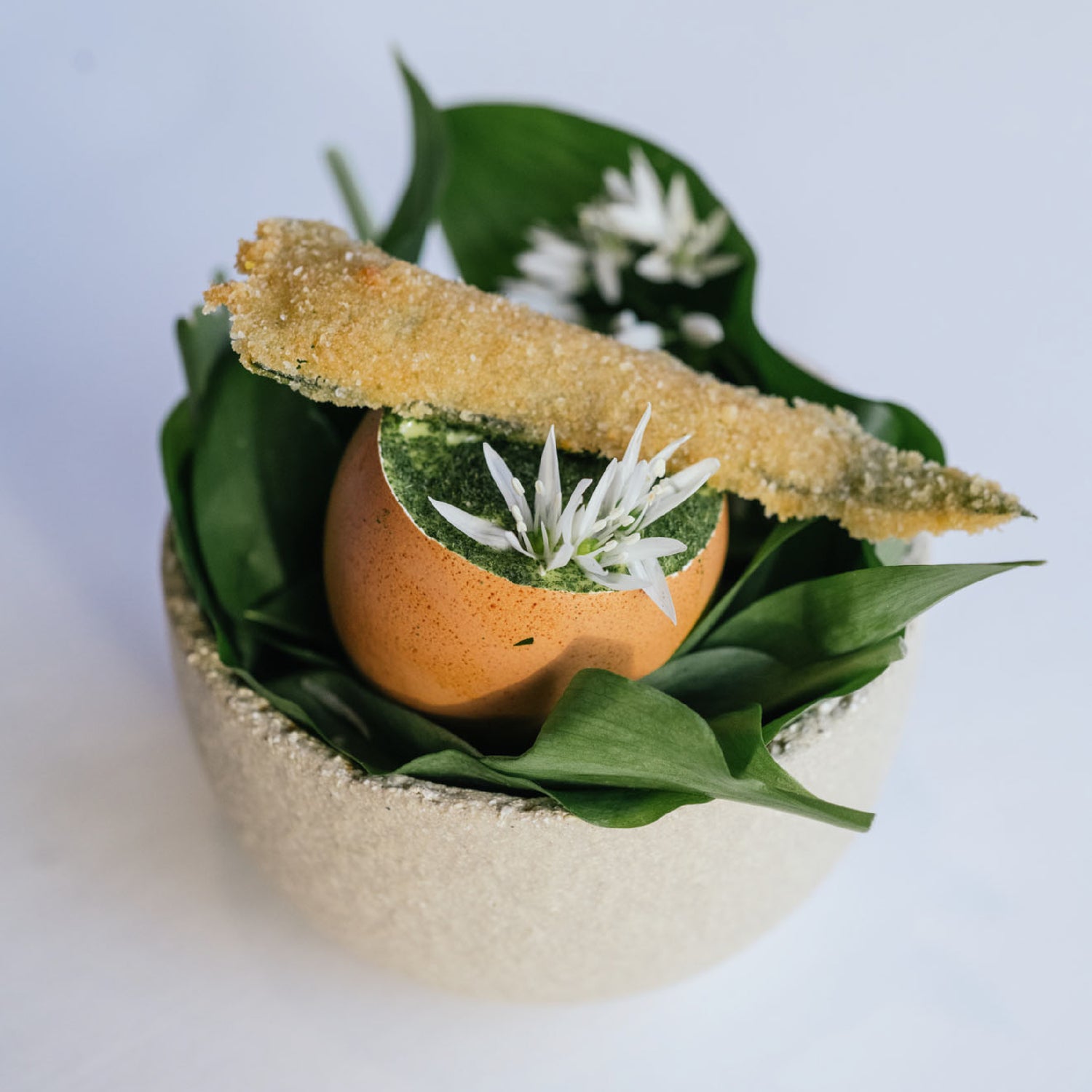 Un œuf servi dans sa coquille, garni de mousse verte et décoré de petites fleurs blanches, accompagné d'un bâtonnet croustillant, le tout présenté sur un lit de feuilles vertes dans un bol élégant.