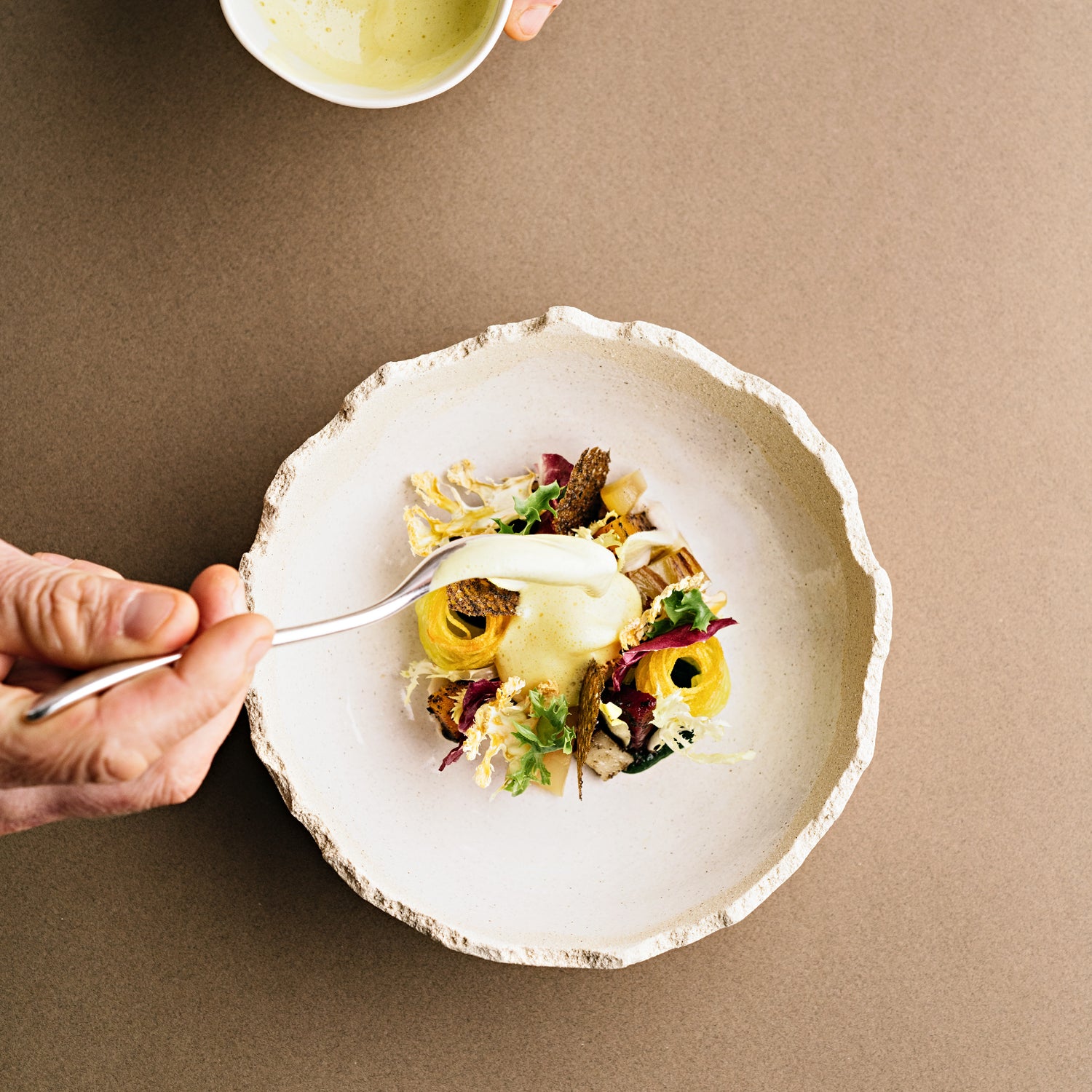 Un plat raffiné dans une assiette en céramique texturée, garni de légumes délicatement disposés, tandis qu'une main ajoute une sauce crémeuse depuis une cuillère, apportant la touche finale à cette présentation gastronomique.