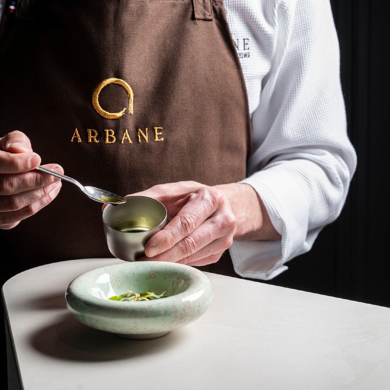 Chef en tablier Arbane préparant un plat délicat avec une petite coupelle et une cuillère au-dessus d’un bol en céramique verte.
