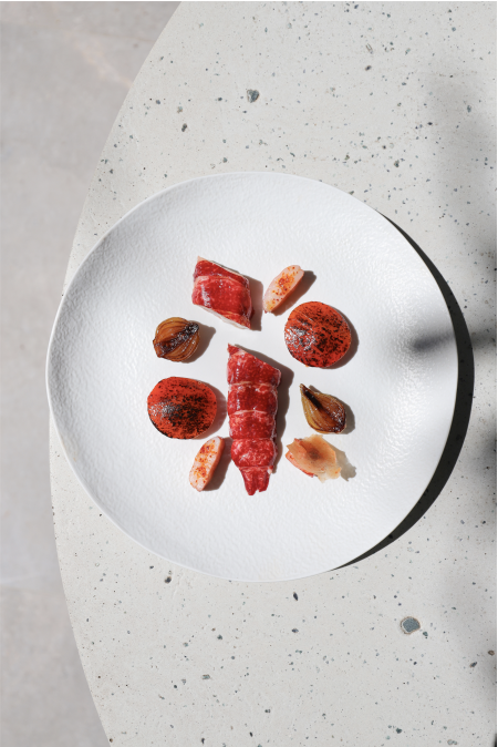 Assiette gastronomique minimaliste avec des morceaux de viande et de légumes grillés, disposés de manière épurée sur une assiette blanche texturée, présentée sur une surface en pierre.