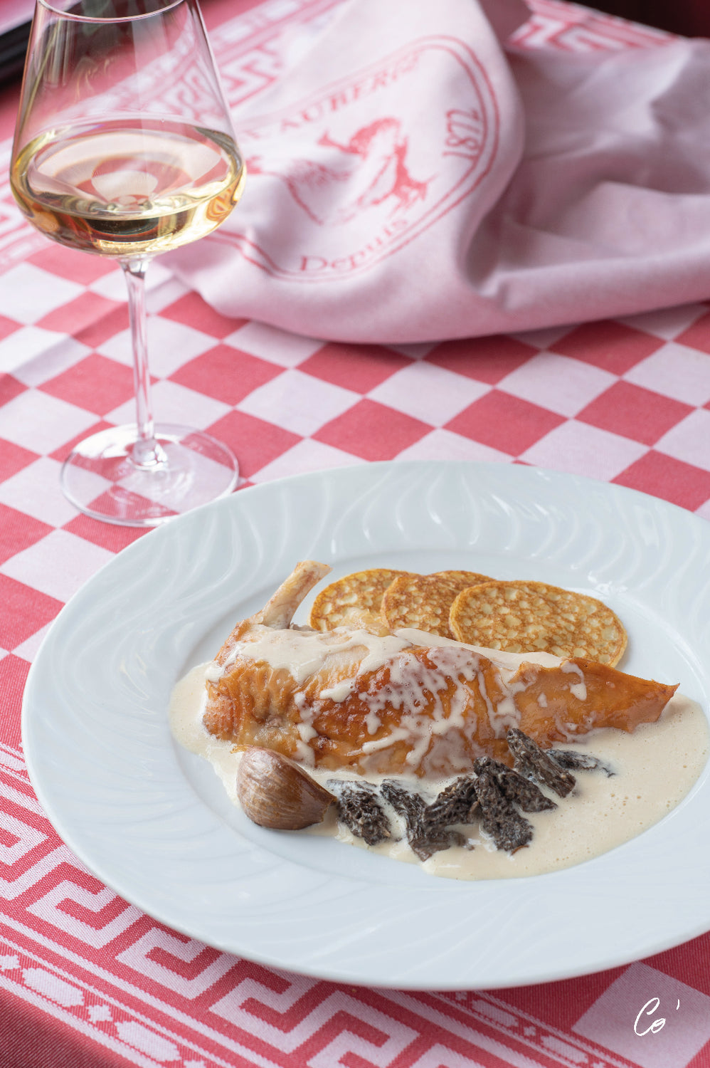 Plat de volaille sauce crème aux morilles, accompagné de galettes, servi sur une assiette blanche avec un verre de vin blanc, sur une nappe à carreaux roses et blancs.