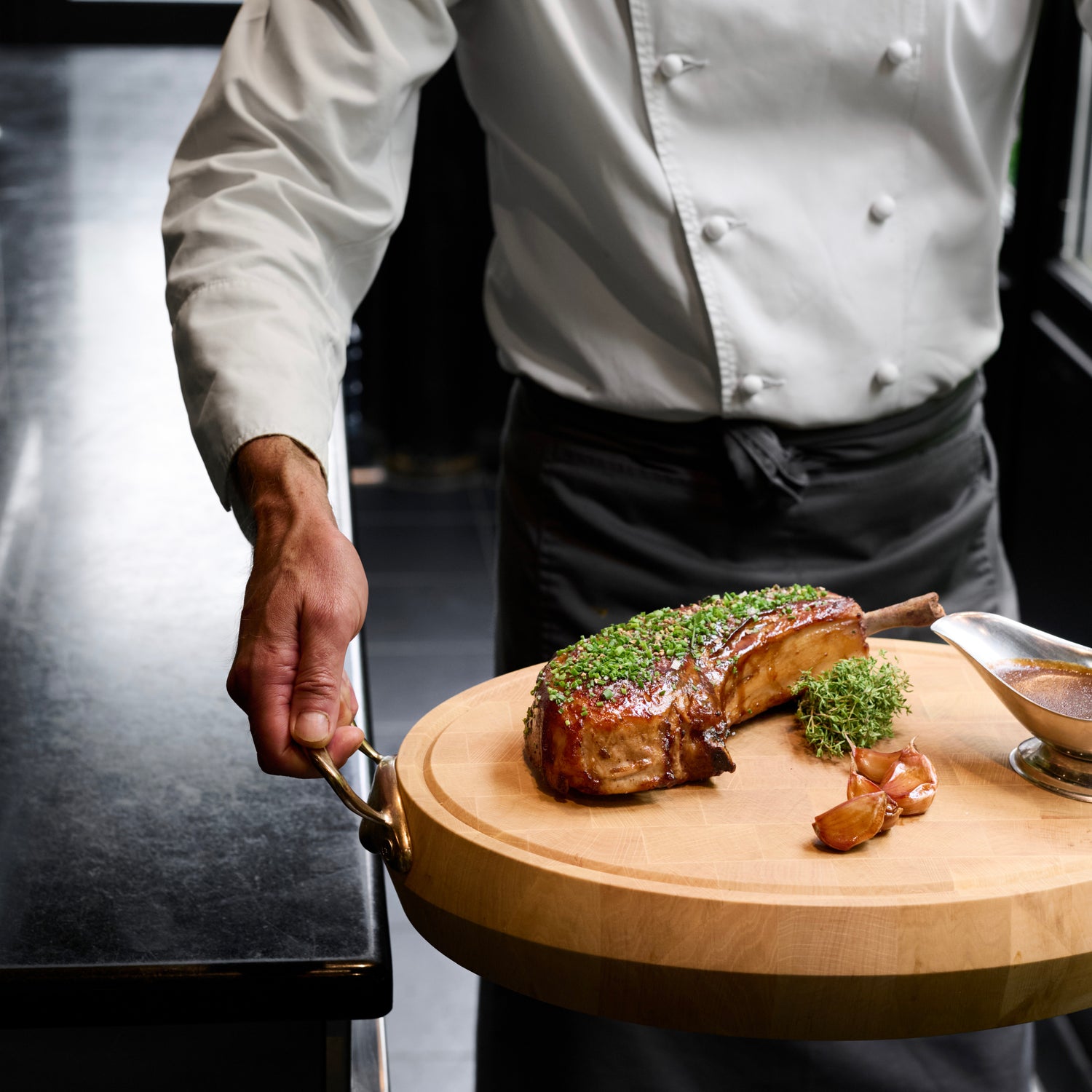 Chef tenant une planche de bois avec une côte de porc garnie d'herbes et des gousses d'ail rôties