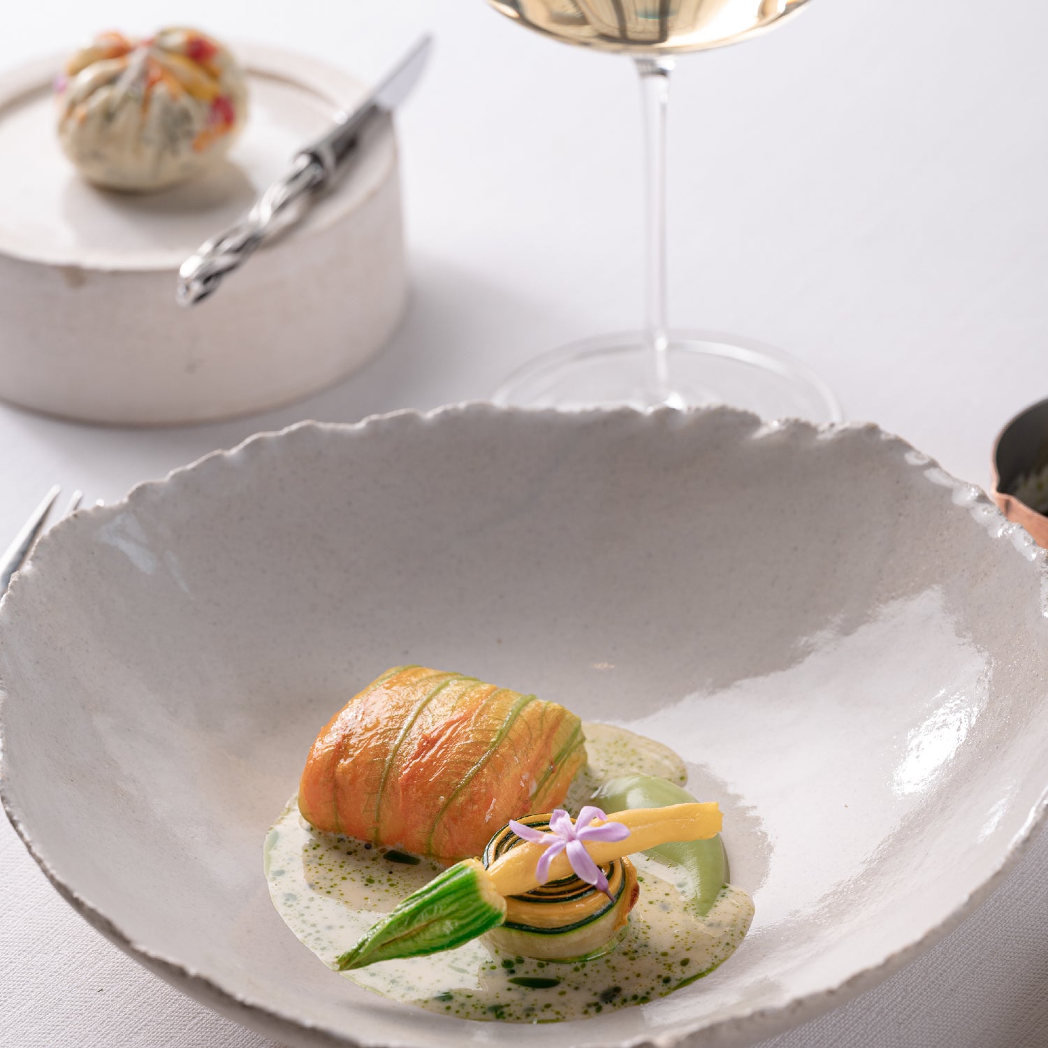 Plat gastronomique composé d'un roulé de légumes délicatement dressé dans une assiette en céramique, accompagné d'une sauce onctueuse et décoré d'une fleur comestible, avec un verre de vin blanc en arrière-plan.