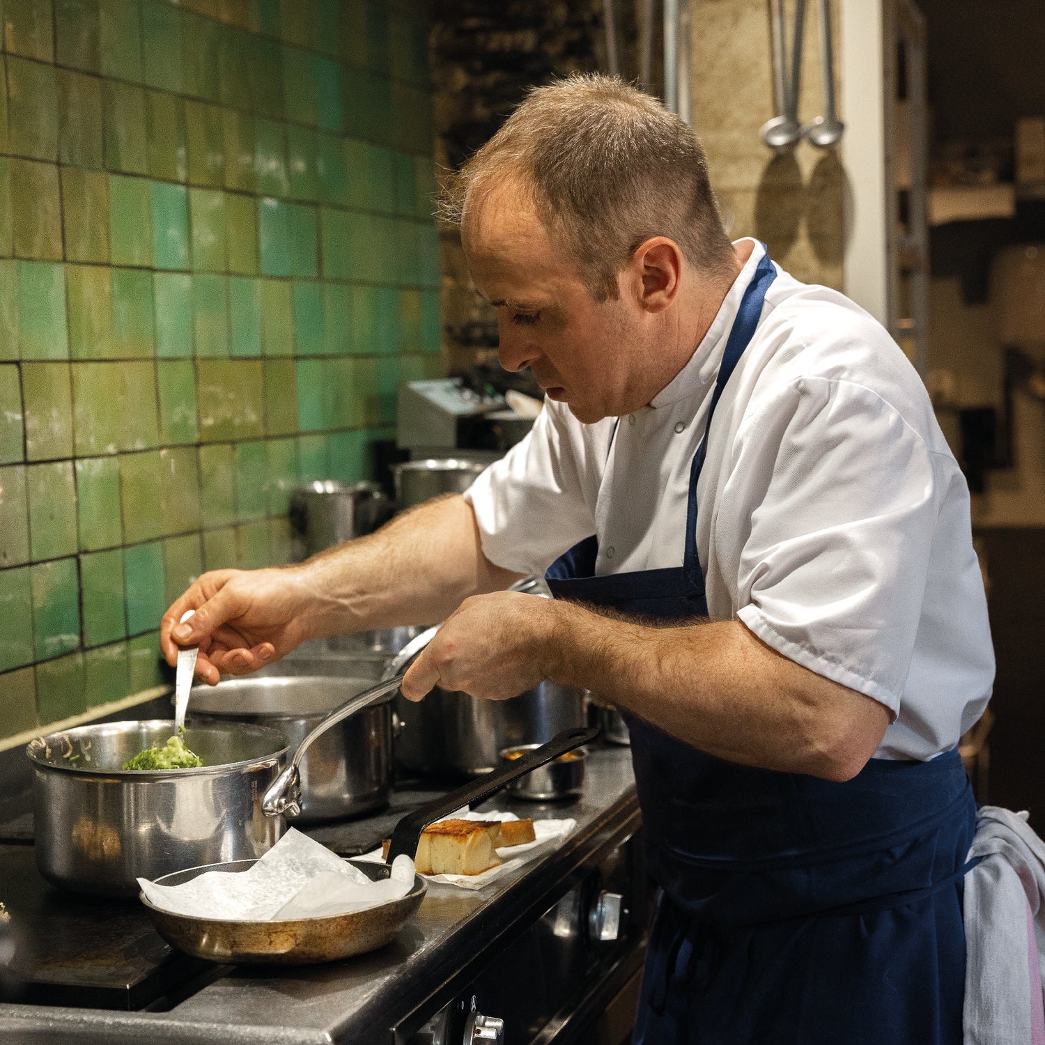 Un chef cuisinier concentré en train de préparer un plat dans une cuisine professionnelle avec des carreaux verts en arrière-plan.