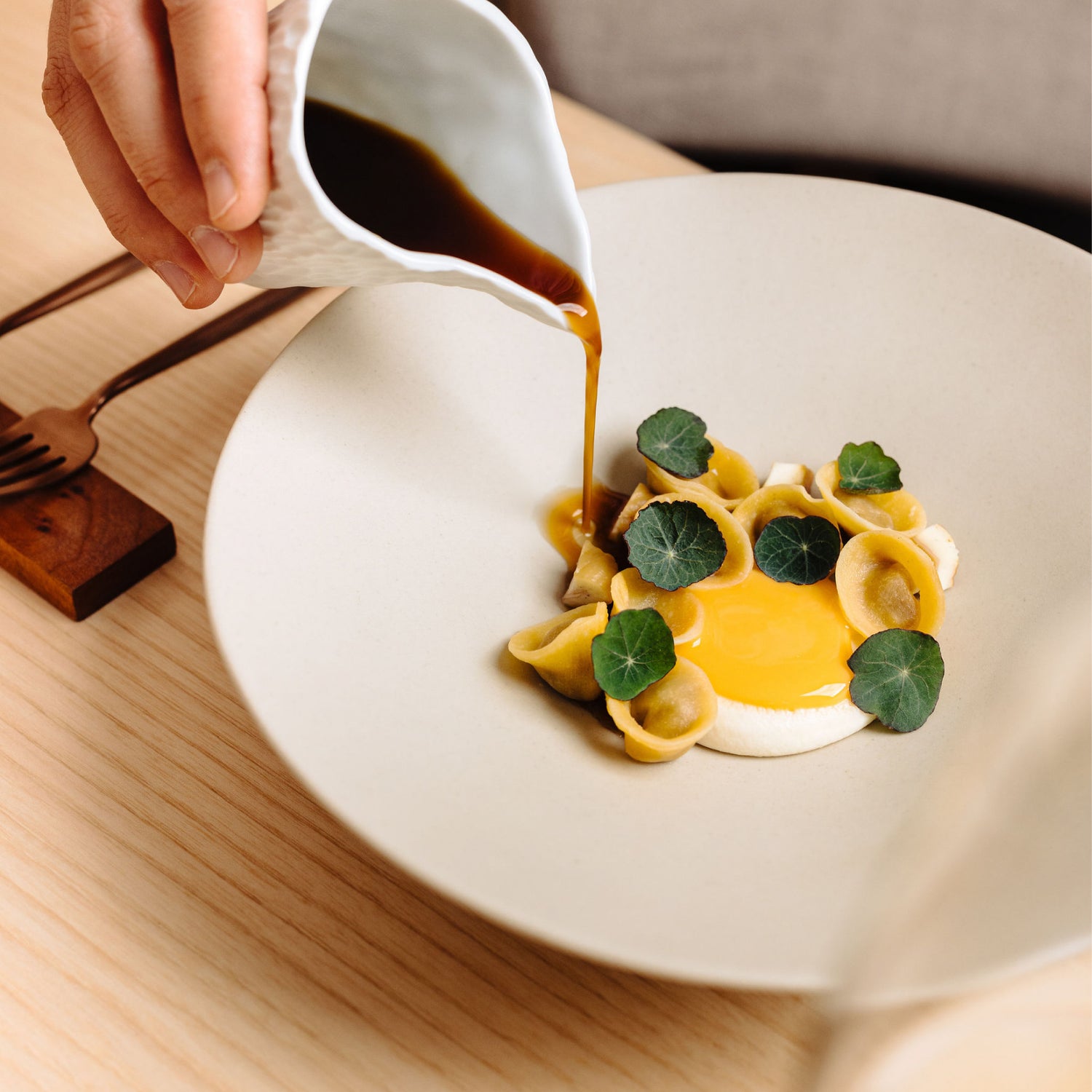 Plat gastronomique de pâtes artisanales garnies de feuilles de capucine, avec une sauce brune versée délicatement depuis une petite carafe blanche, présenté sur une table en bois clair.