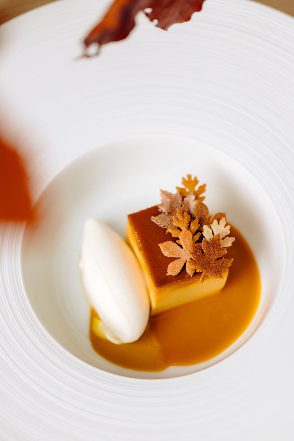 Dessert raffiné composé d'un flan caramel accompagné de tuiles croquantes en forme de feuilles, servi avec une quenelle de glace vanille sur un lit de sauce onctueuse, présenté dans une assiette blanche élégante.