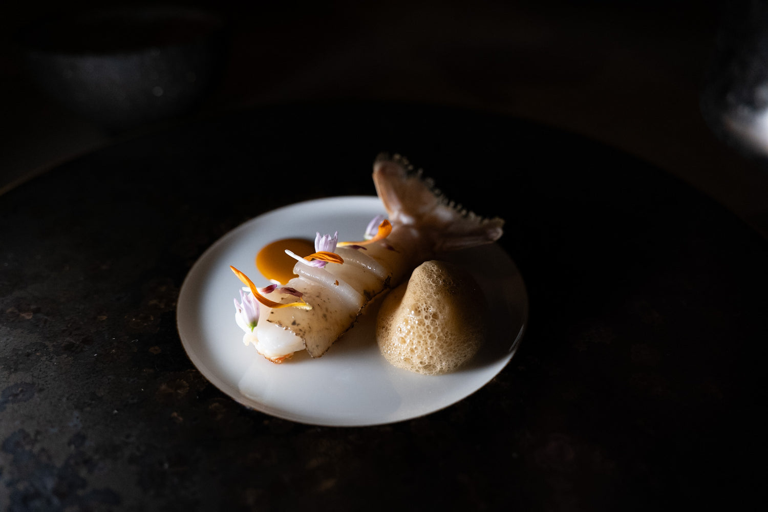 Plat gastronomique raffiné composé de tranches de poisson blanc délicatement disposées avec des fleurs comestibles sur une assiette blanche, accompagné d'une sauce et d'une mousse légère, dans une ambiance feutrée et élégante.