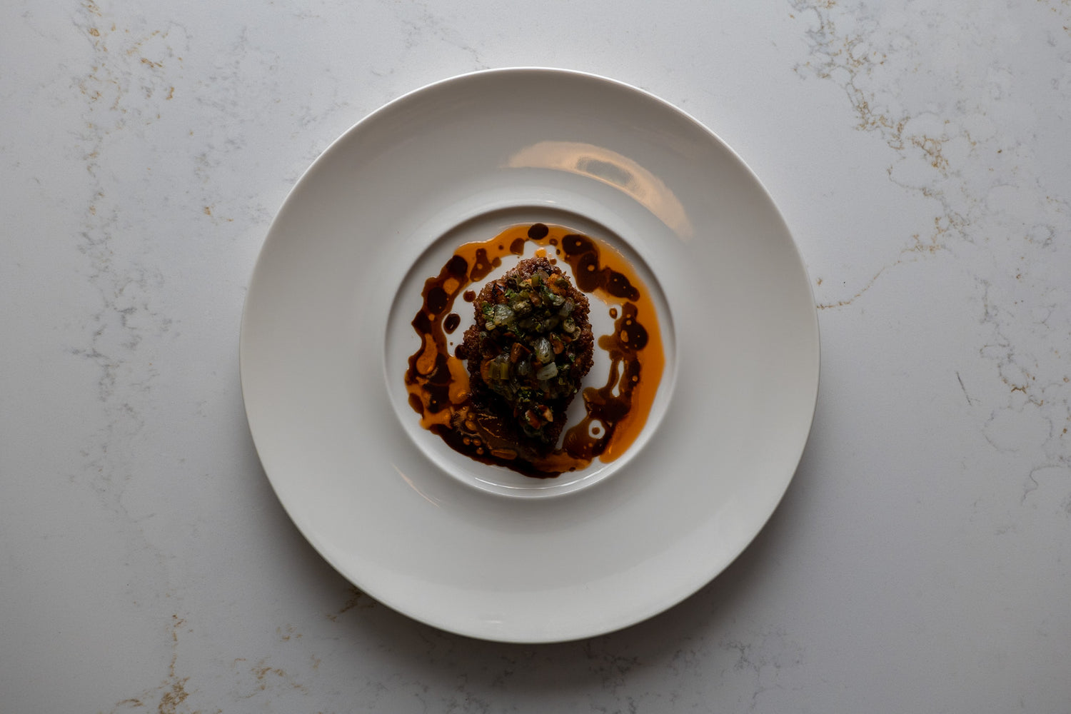 Une assiette gastronomique vue de dessus, présentant un plat délicatement dressé au centre. Le mets est entouré d'une sauce brun foncé, disposée de manière artistique sur l'assiette blanche, créant un contraste avec la couleur claire de l'assiette et de la surface marbrée.