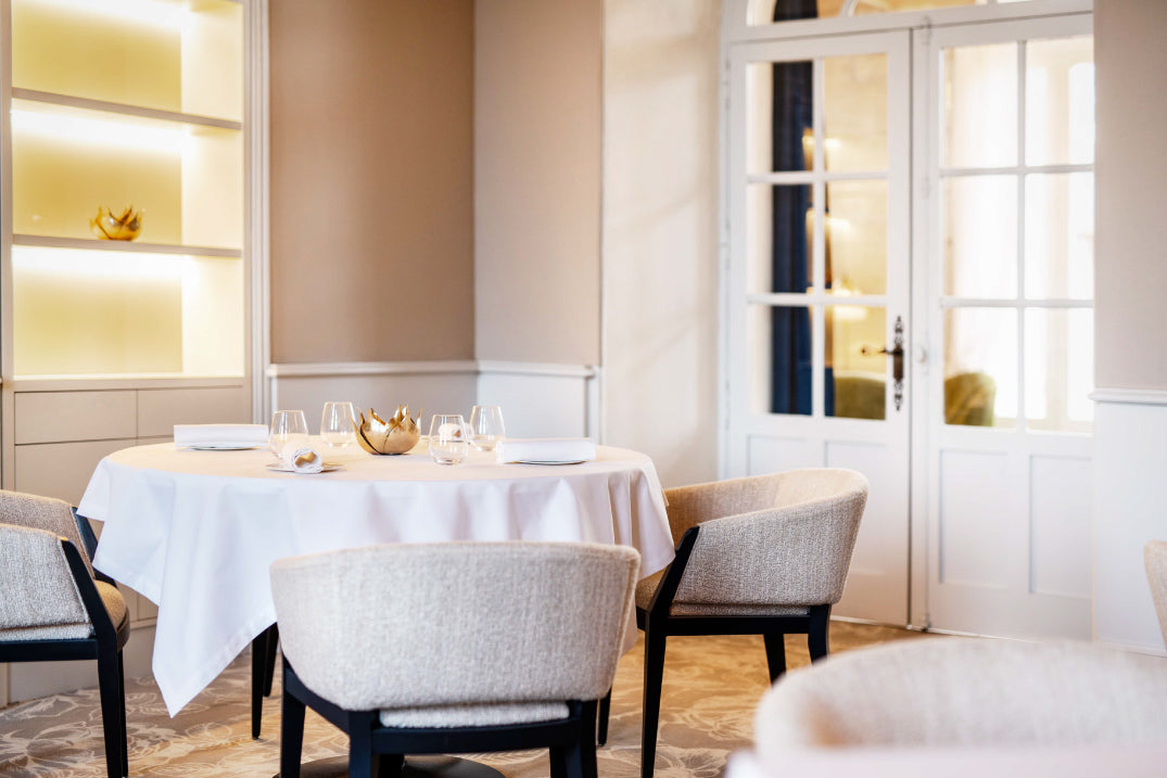 Table ronde élégamment dressée avec une nappe blanche, entourée de chaises rembourrées dans une salle de restaurant lumineuse, avec des étagères rétroéclairées et des portes vitrées donnant sur une autre pièce.