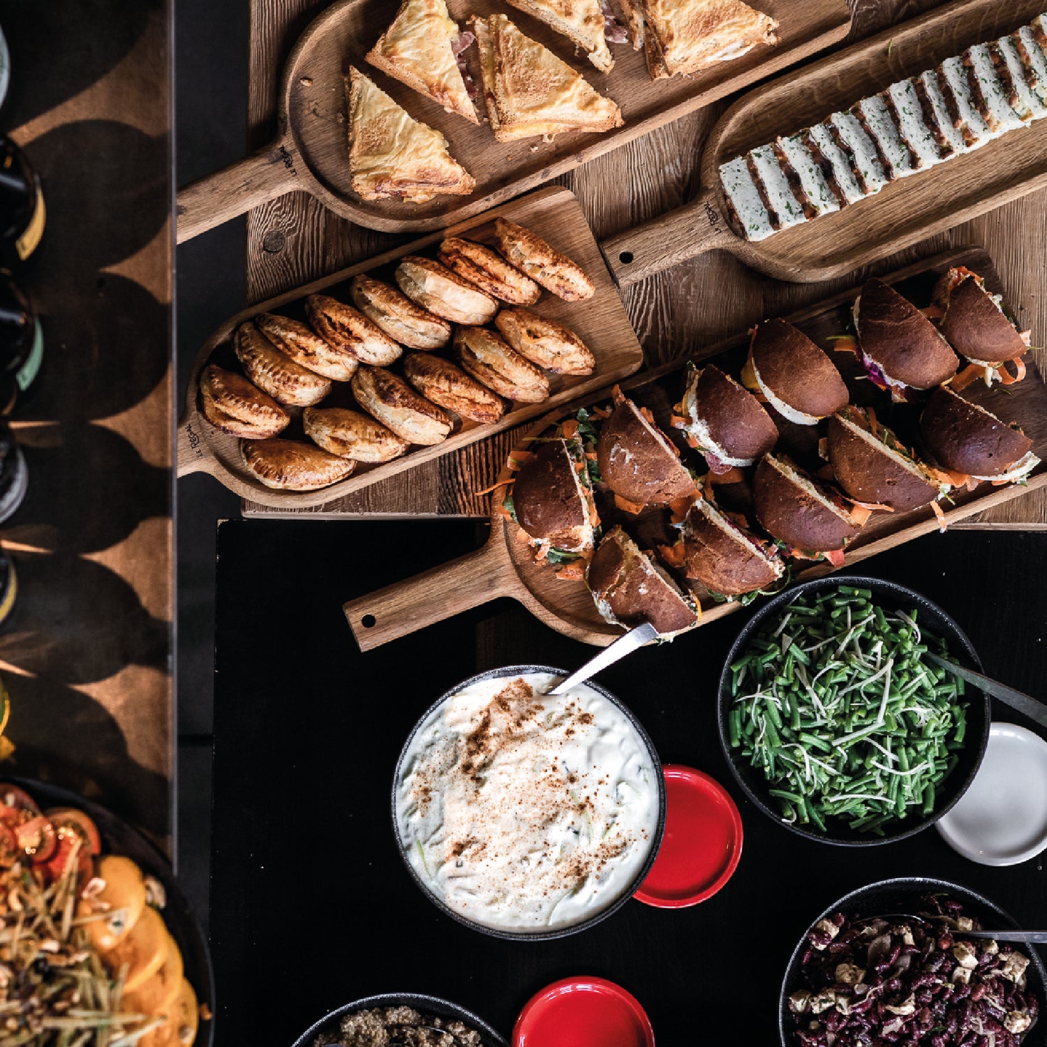 Vue aérienne d'un buffet varié comprenant des sandwichs, des feuilletés, des pâtisseries salées et des bols de salades et de sauces, disposés sur des planches en bois.