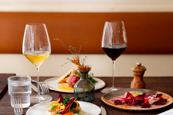 Table de restaurant élégamment dressée avec deux verres de vin (blanc et rouge), des assiettes de plats raffinés colorés, un verre d’eau, une salière en bois et un petit vase décoratif au centre.