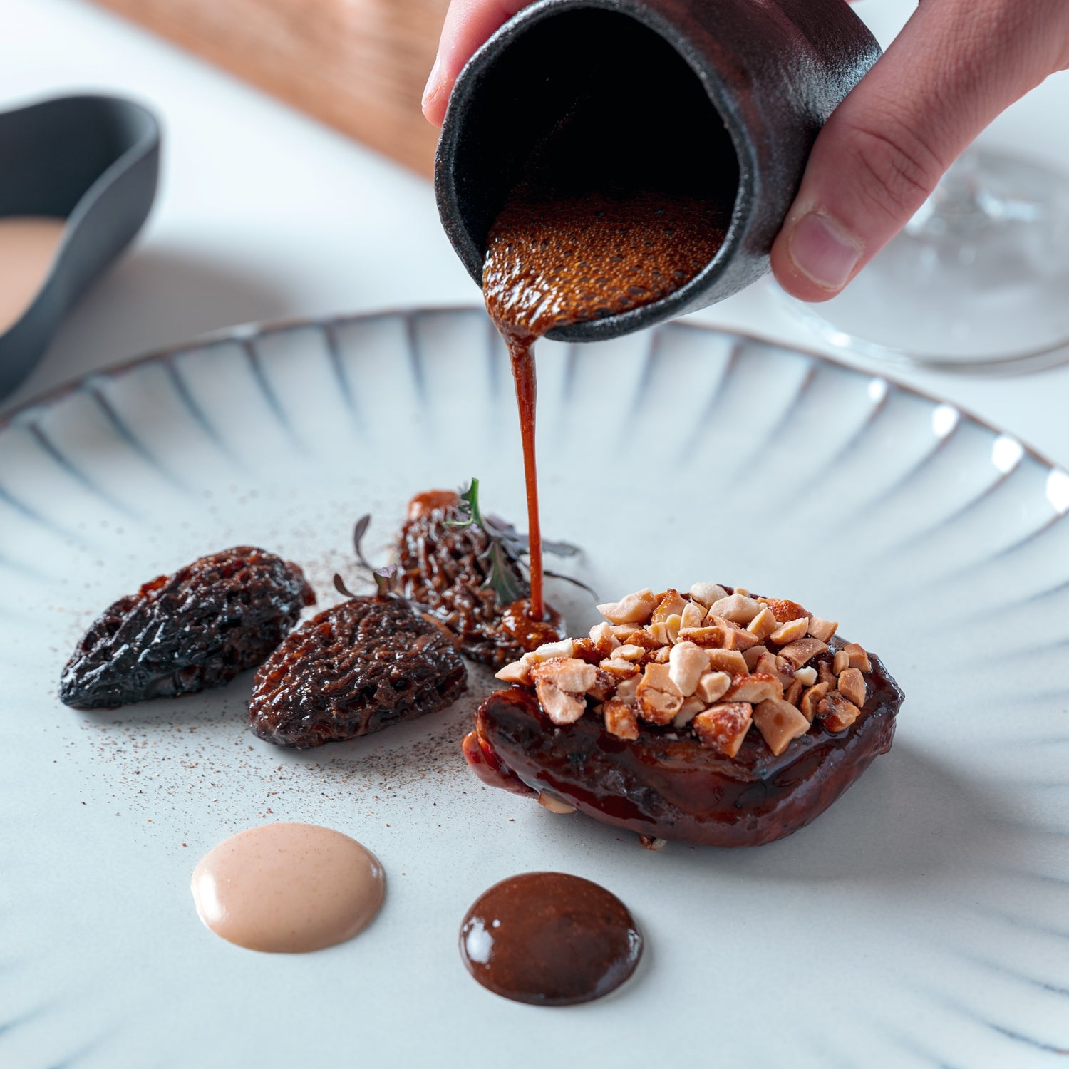 Un plat gastronomique composé d'un morceau de viande recouvert de cacahuètes concassées, accompagné de morilles et de deux sauces en gouttes, sur lequel une sauce riche est versée délicatement à partir d'une petite cruche noire.