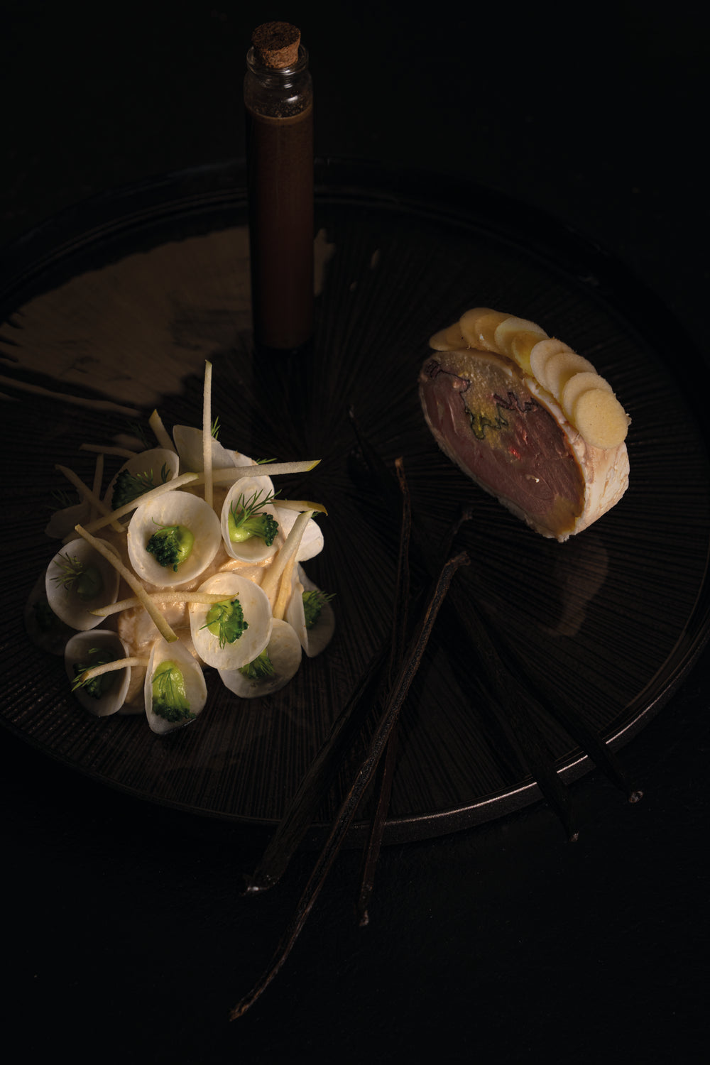 Plat gastronomique composé de légumes finement tranchés et disposés avec élégance à côté d'une pièce de viande garnie de fines lamelles, le tout présenté sur une assiette sombre et décoré de manière artistique, mettant en valeur l'aspect sophistiqué du mets.