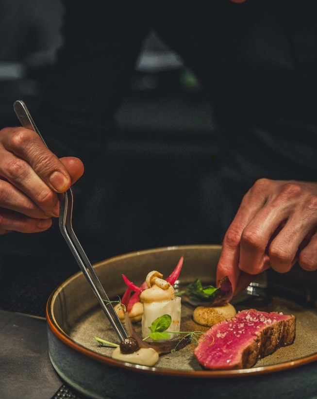 Chef ajustant une assiette de viande et de légumes finement décorée avec des pinces.