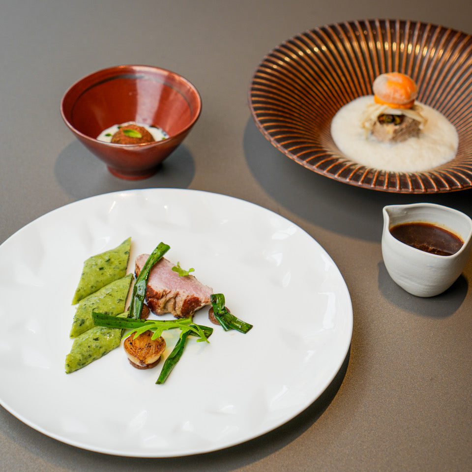 Assortiment de plats gastronomiques comprenant une assiette de viande avec légumes et sauce, un bol de sauce à part, et deux bols contenant des préparations créatives, présentés sur une table.