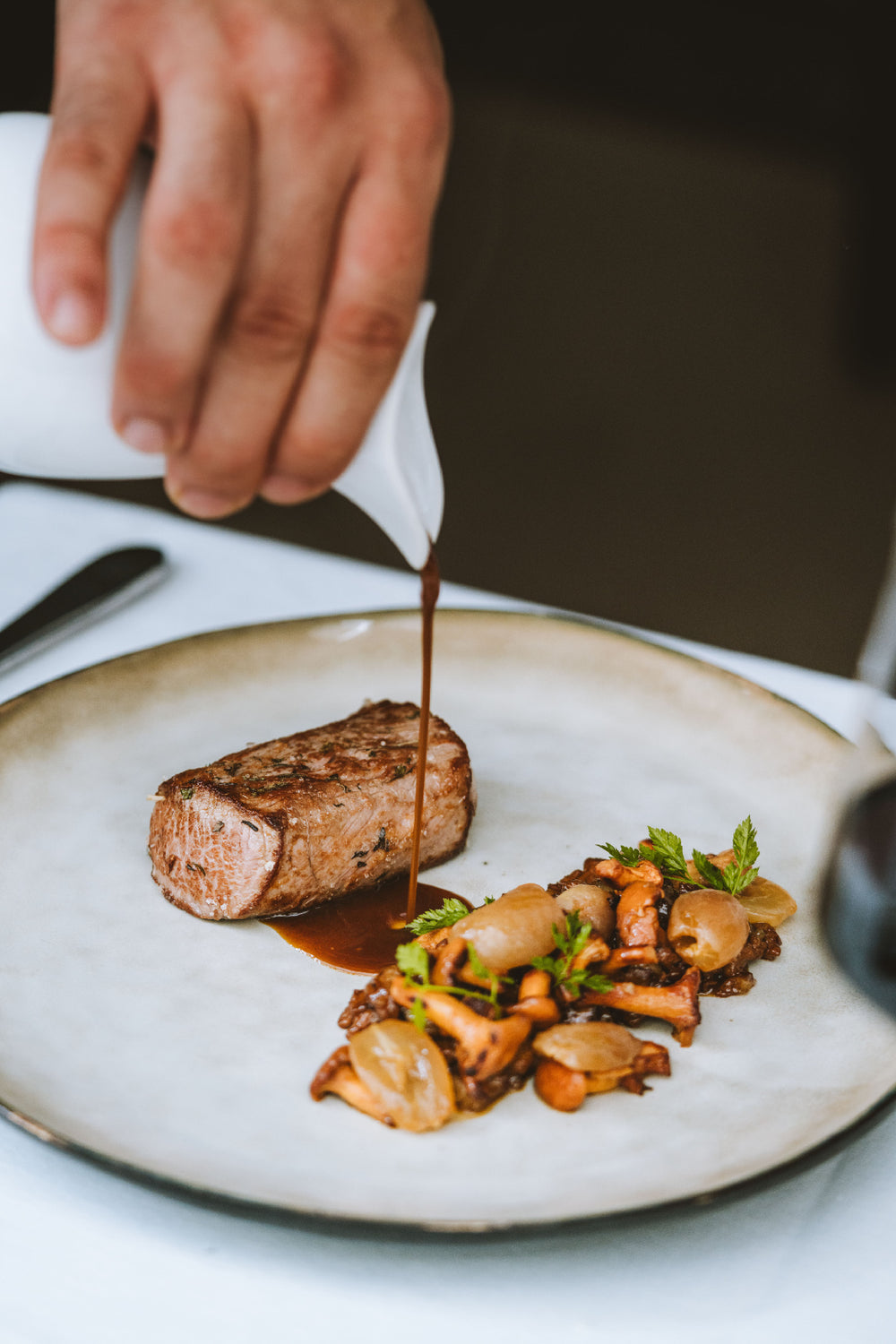 Main de chef versant délicatement une sauce sur un morceau de viande grillée, accompagnée de champignons et d'oignons caramélisés sur une assiette élégante.