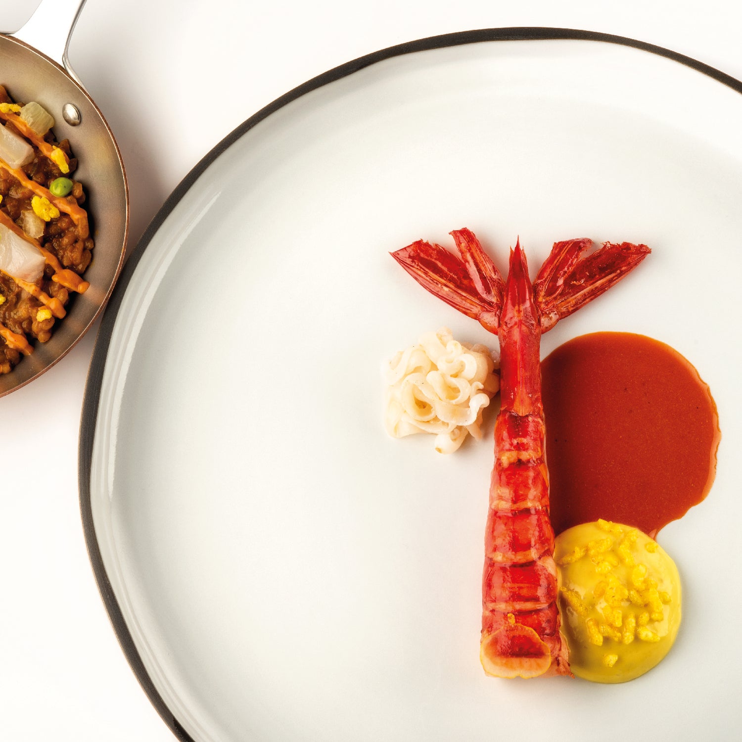 Assiette gastronomique composée d'une crevette rouge dressée avec une sauce brune, une mousse jaune, et des nouilles, accompagnée d'une poêle de paëlla garnie de légumes et de poisson.