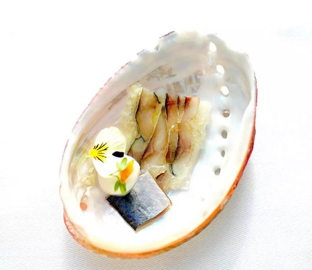 Un plat raffiné présenté dans une coquille, avec des tranches délicates de poisson cru, accompagné de fleurs comestibles et d'une garniture élégante, le tout sur un fond blanc épuré.