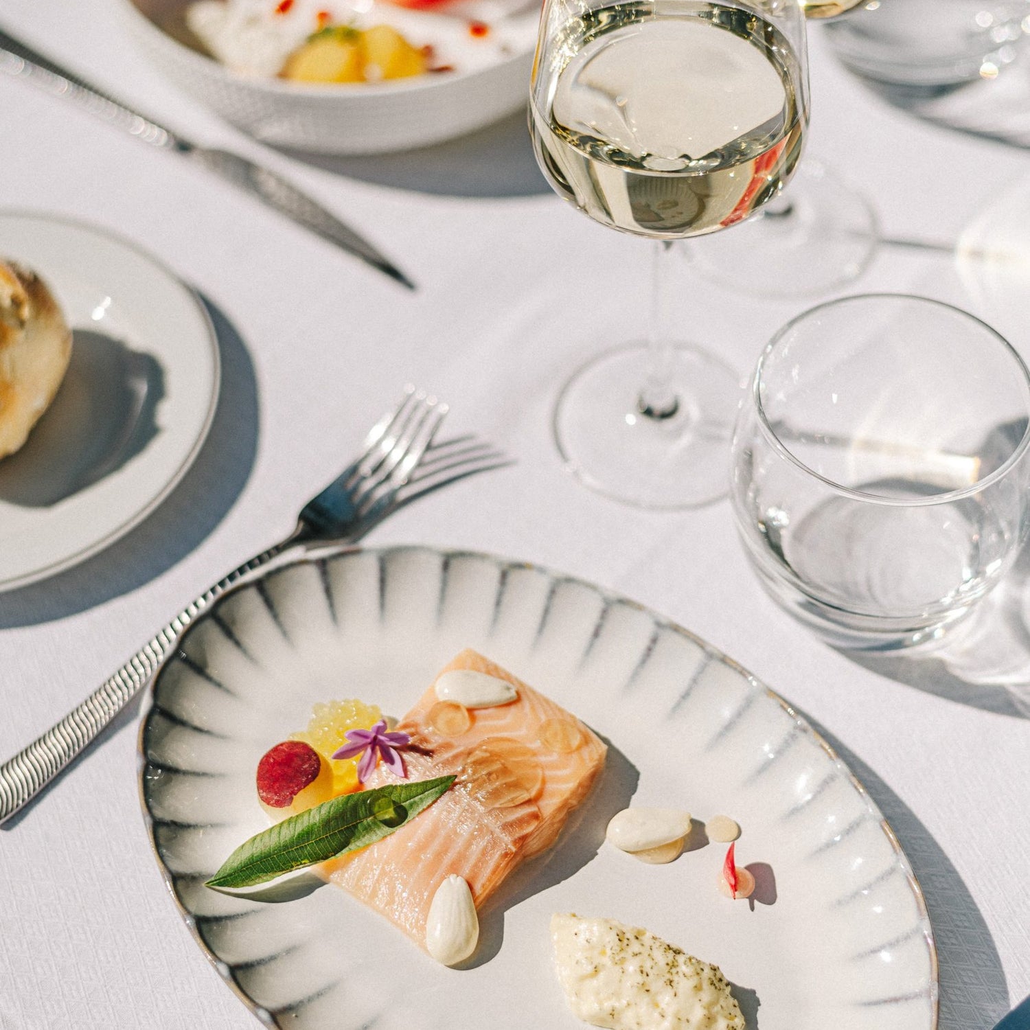 Table dressée sur une nappe blanche, photo prise à l'extérieur sous un beau soleil. Dans les assiettes des plats raffinés. Deux verres de vin blanc devant les assiettes.