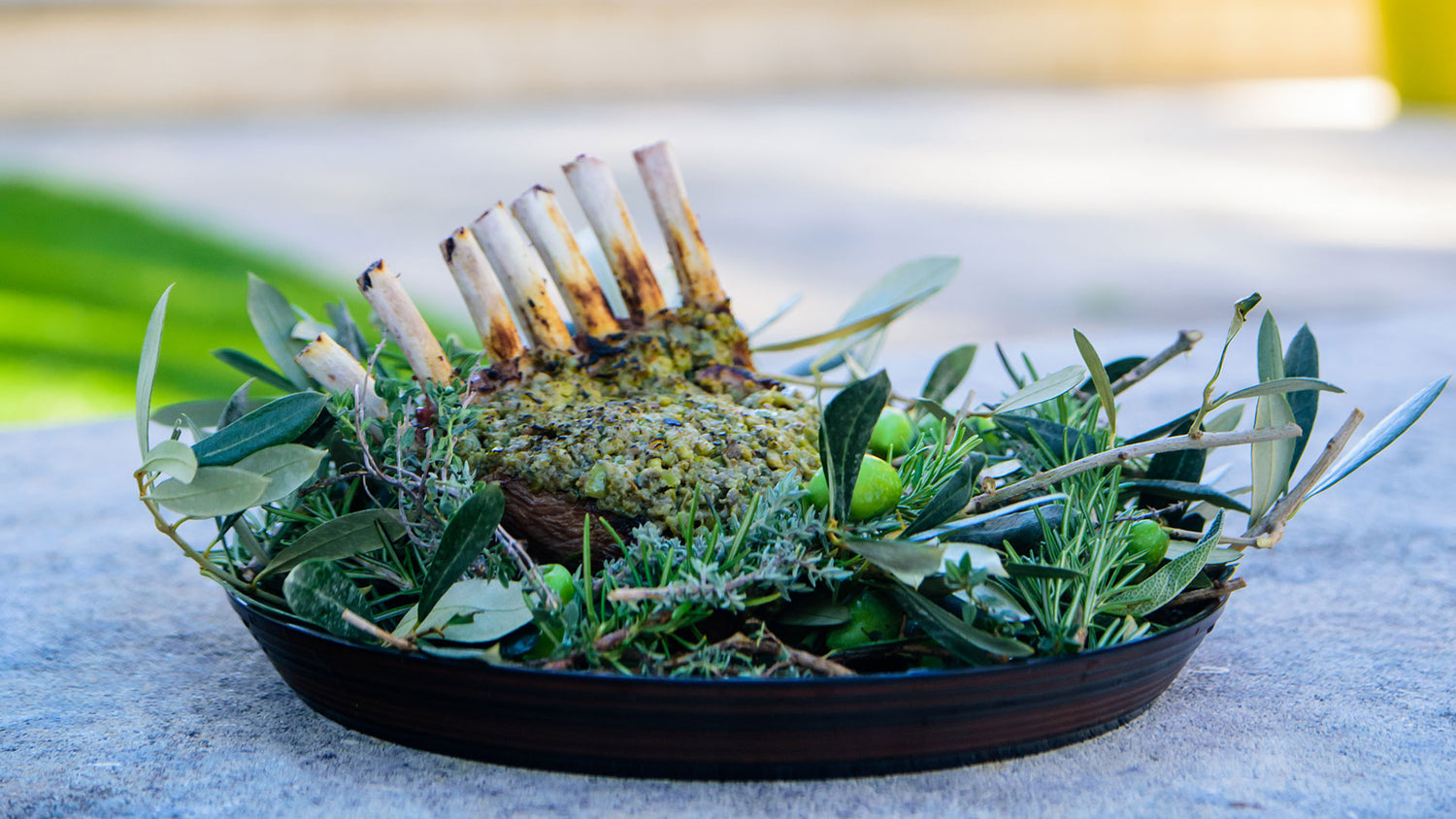 Carré d'agneau rôti, servi sur un lit de branches d'herbes fraîches, présenté sur une assiette rustique en plein air.