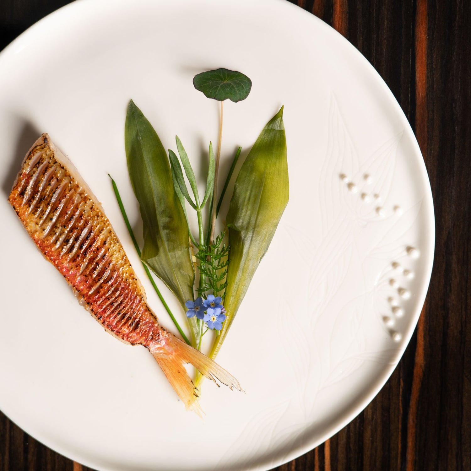 Assiette minimaliste présentant un filet de poisson grillé accompagné de jeunes pousses et de fleurs comestibles.