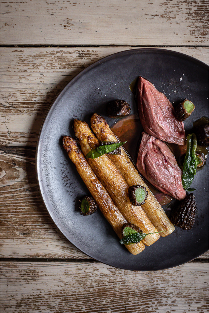 Assiette gastronomique avec des asperges rôties, accompagnées de morceaux de viande rosée et de morilles, présentée sur une assiette noire et posée sur une table en bois rustique.