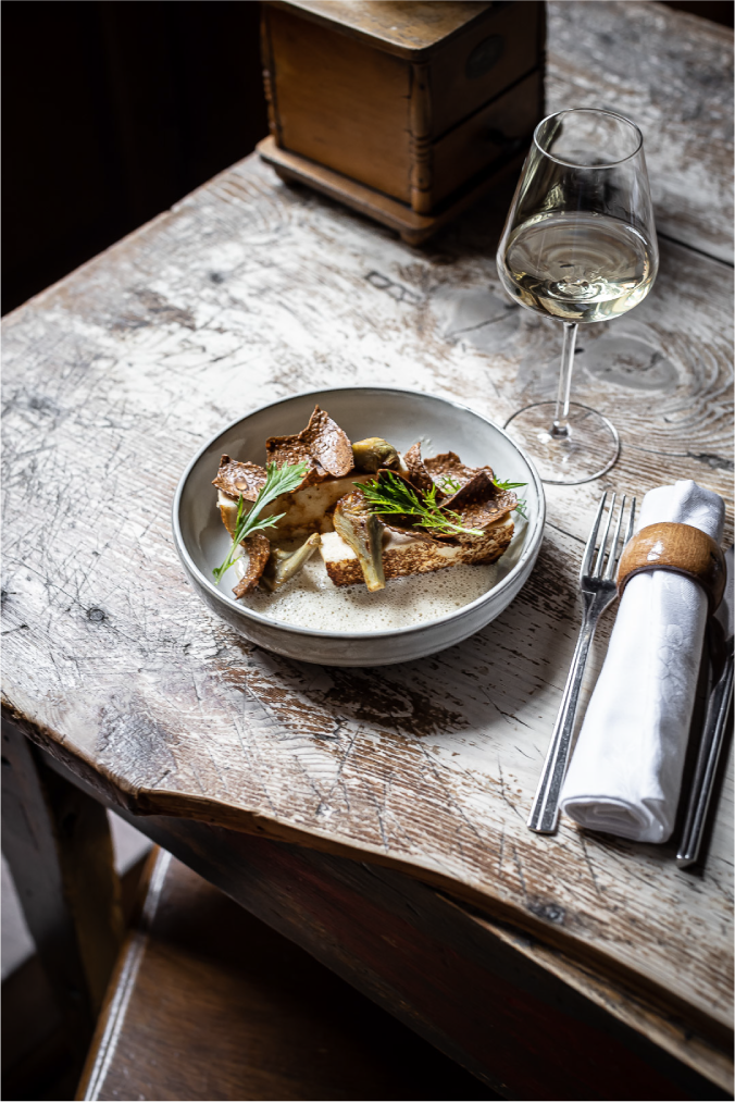 Plat gastronomique servi dans une assiette creuse sur une table en bois rustique, accompagné d'un verre de vin blanc. Le plat est composé de légumes rôtis garnis d'herbes fraîches, avec un moulin à café vintage en arrière-plan.