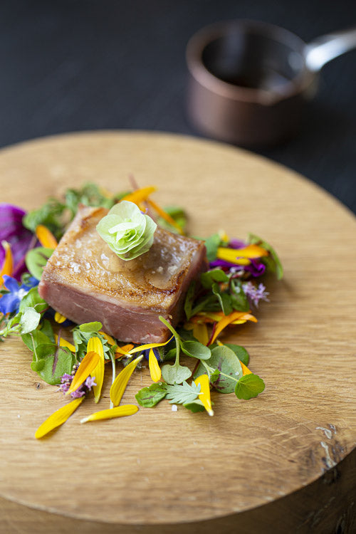 Plat gastronomique élégant composé d'un morceau de viande parfaitement cuit, servi sur un lit de jeunes pousses et de fleurs comestibles, posé sur une planche en bois.