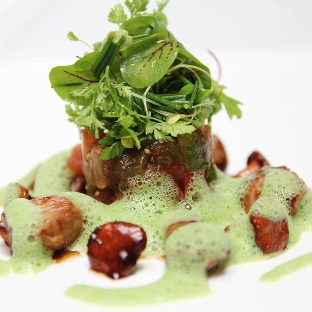 Une assiette contenant un plat savamment dressé, composé de légumes, de champignons, et d'une garniture verte mousseuse. La présentation est artistique, avec un équilibre subtil entre les couleurs et les textures.