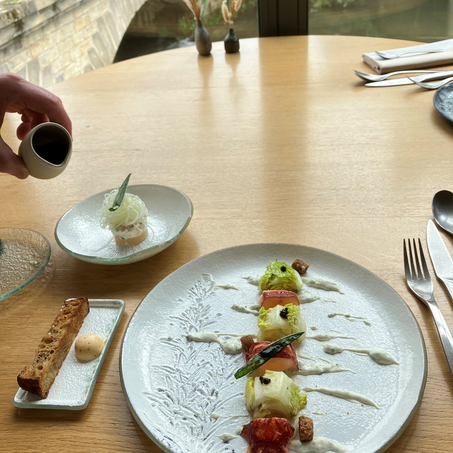 Assiette gastronomique élégante avec des morceaux de homard et de laitue, joliment disposés avec une sauce légère, accompagnée de pain grillé et d'une vue sur un pont et la nature en arrière-plan.