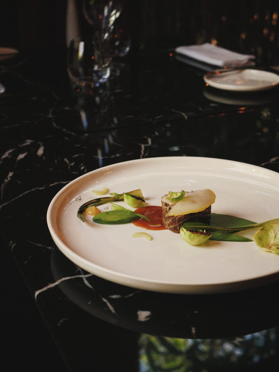 Plat gastronomique soigneusement dressé avec un morceau de poisson rôti, des feuilles vertes et des touches de sauce sur une assiette blanche, posé sur une table en marbre noir élégant.