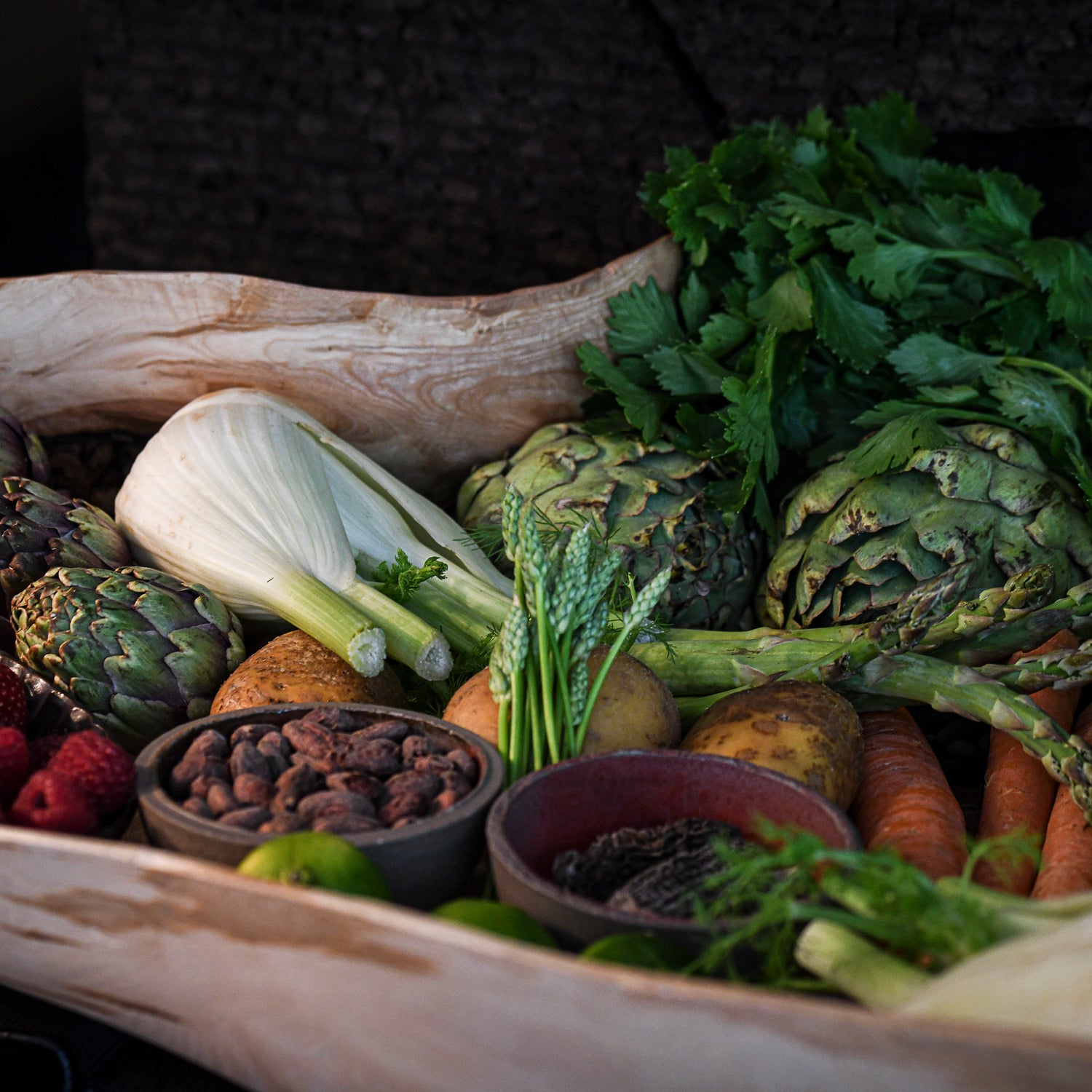 Un panier en bois rempli de légumes frais, dont des artichauts, du fenouil, des asperges, des carottes, des pommes de terre et des céleris, ainsi que des fraises et des framboises.