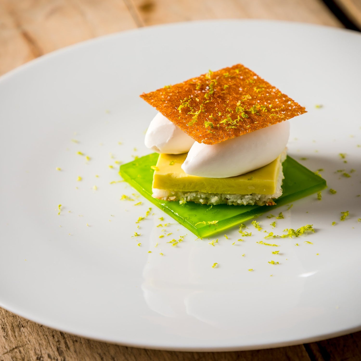 Un dessert sophistiqué avec une présentation en couches, surmonté d'une fine croustille et de zeste de citron vert, servi sur une assiette blanche.
