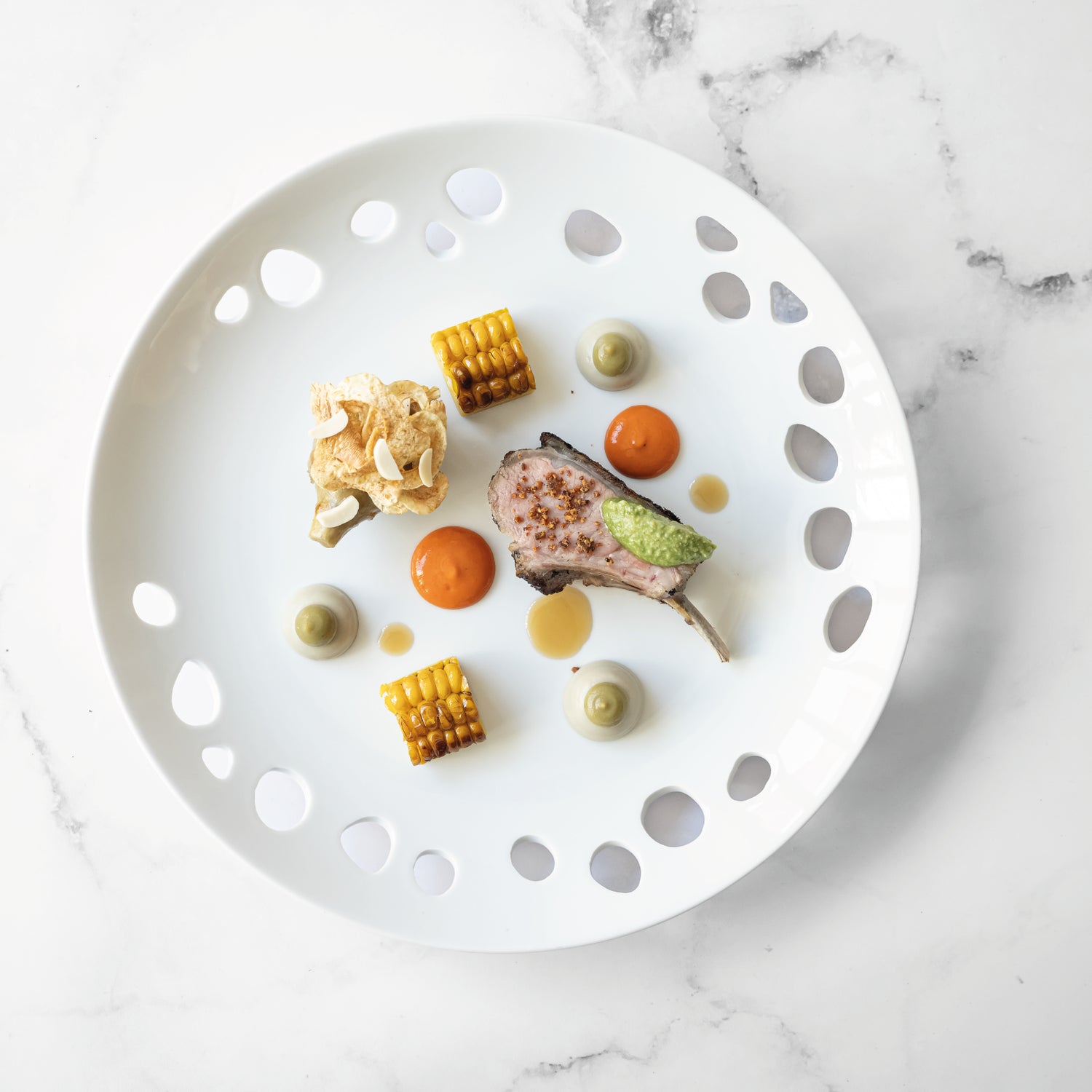 Assiette gastronomique élégamment dressée avec un carré d'agneau, des cubes de maïs grillé, des purées colorées et des croustillants, sur une assiette blanche ajourée.