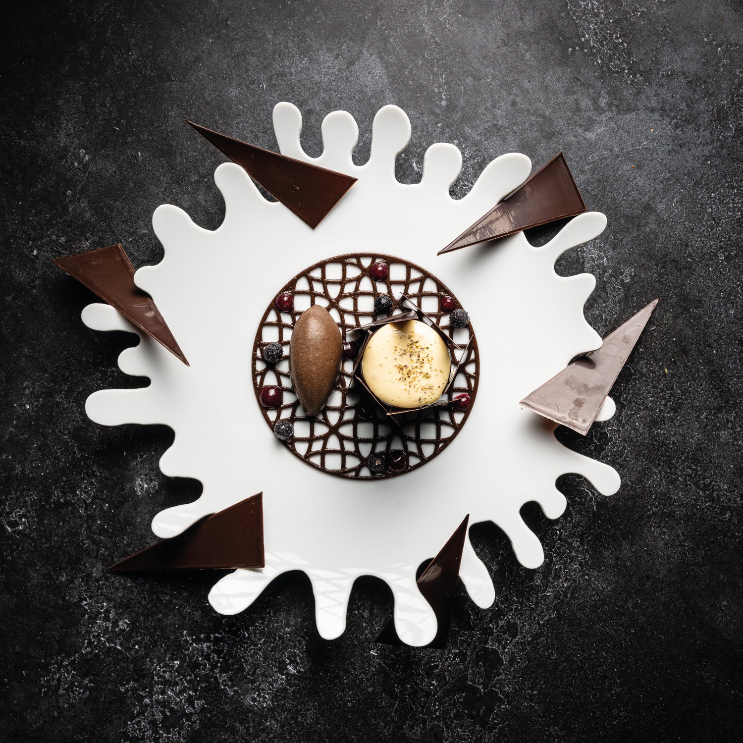 "Dessert gastronomique artistique composé de mousse au chocolat, d'une glace et de décorations en chocolat, présenté sur une assiette blanche éclaboussée de motifs abstraits, le tout sur une table sombre élégante.