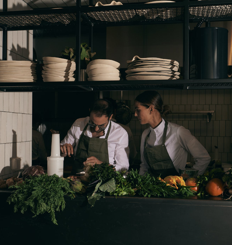 Deux chefs en cuisine préparant des plats, entourés de légumes et herbes fraîches
