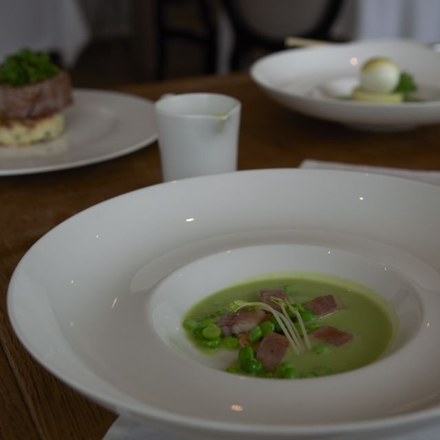 Photo d'un bol de soupe verte, décoré avec des morceaux de viande et de légumes verts, avec un plat de viande en arrière-plan.
