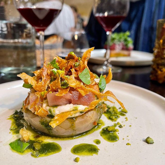 Un plat de tartare de poisson, garni de chips croustillantes et accompagné d'une sauce verte, avec des verres de vin en arrière-plan.